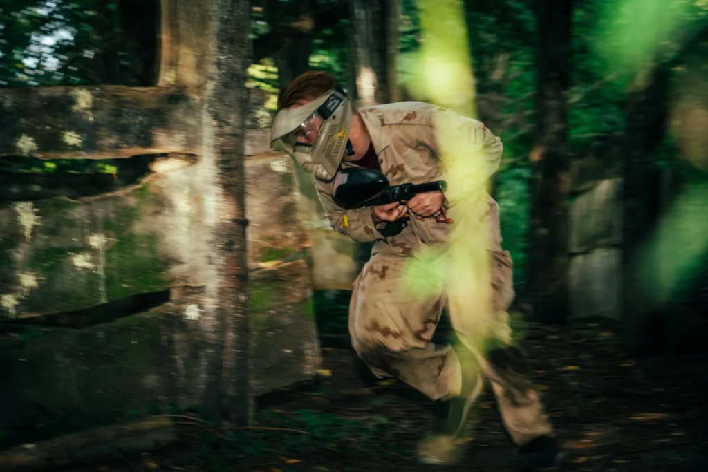 Paintballspeler in camouflagepak sprint met zijn marker langs een houten bunker vol verfspatten
