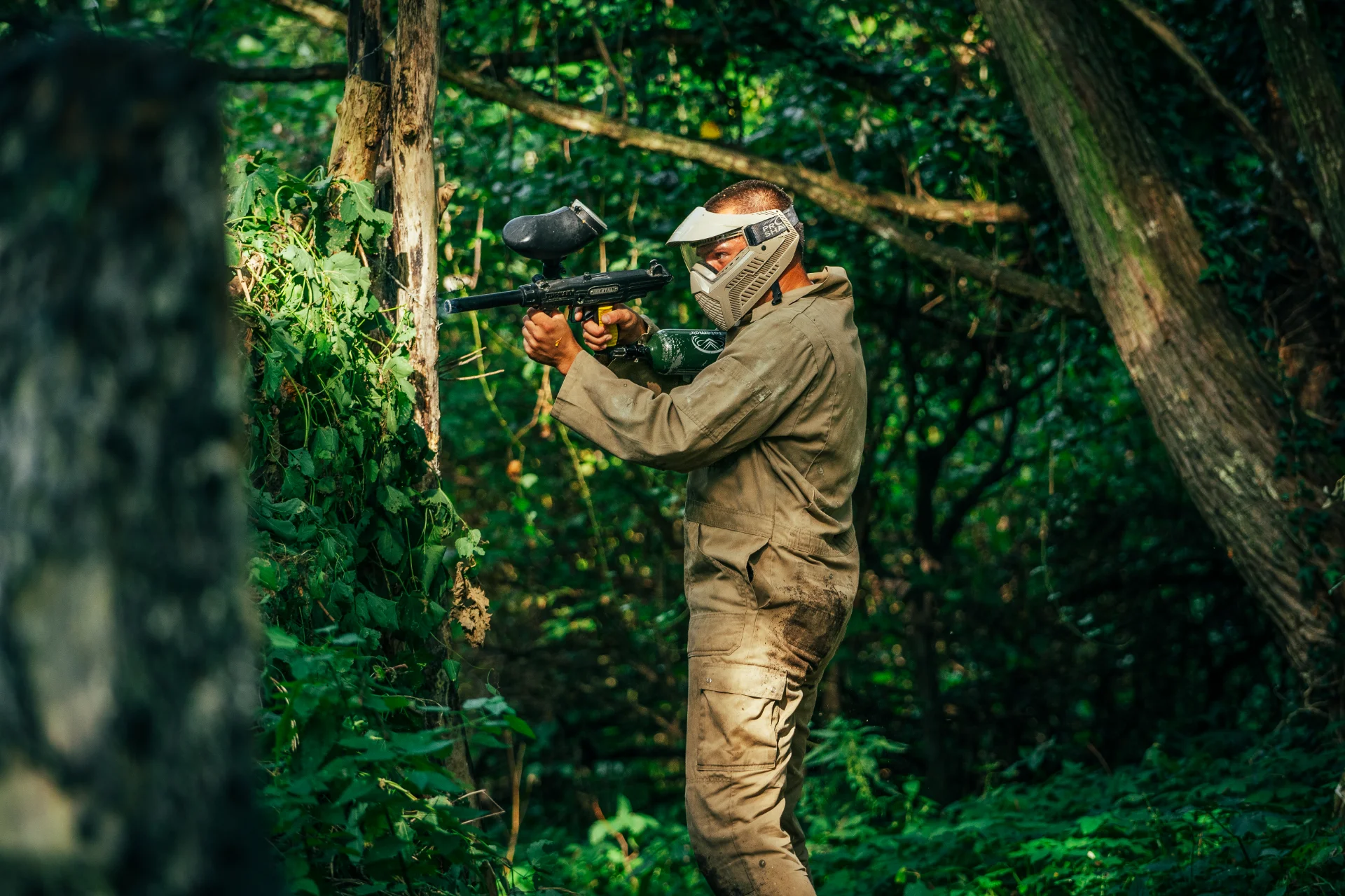 Paintballer in beige overall mikt met zijn marker tussen de bomen tijdens paintball vanuit Markelo